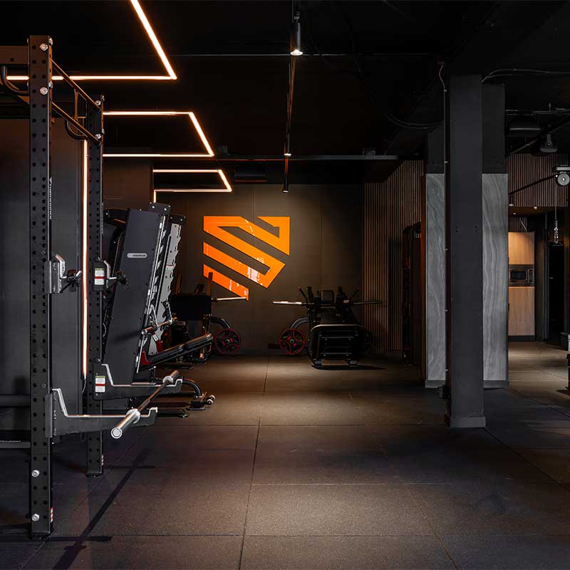 Donkere sportschool met squatracks aan de linkermuur en sportieve ledlijnen. Een oranje logo tegen de achterste wand.